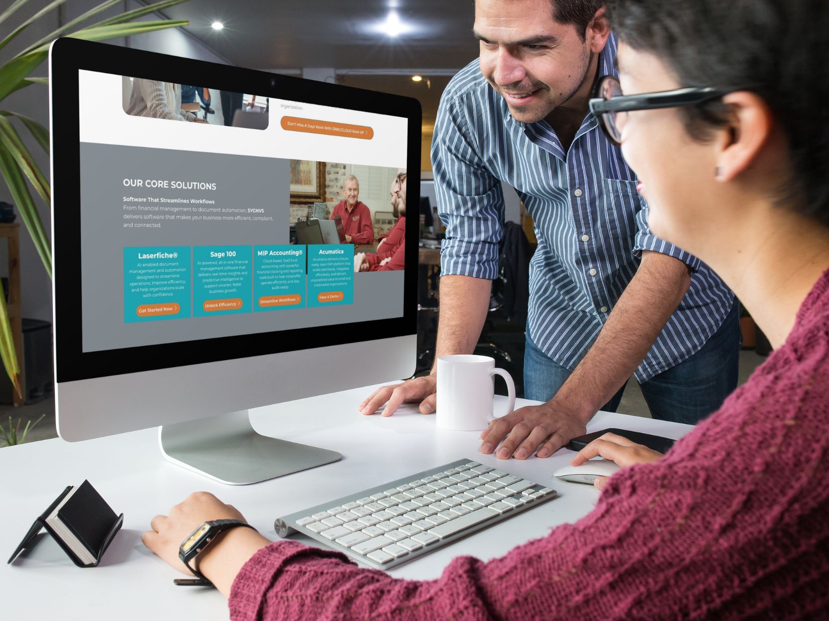 Two coworkers review a project webpage on a large desktop monitor as they lean in and discuss.